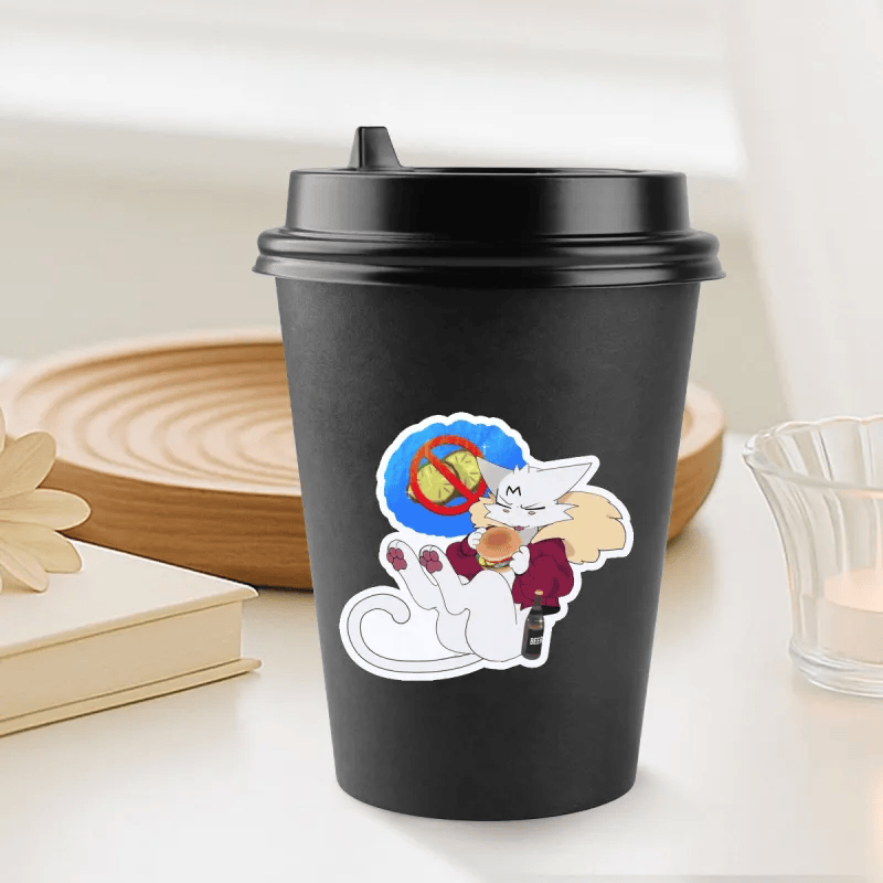 A black reusable coffee cup with a lid sits on a tabletop in a softly styled indoor setting. A die-cut cartoon sticker on the front shows a white cat wearing a red sweater and holding a hot drink, with a blue circular background and a crossed-out hot air balloon symbol.